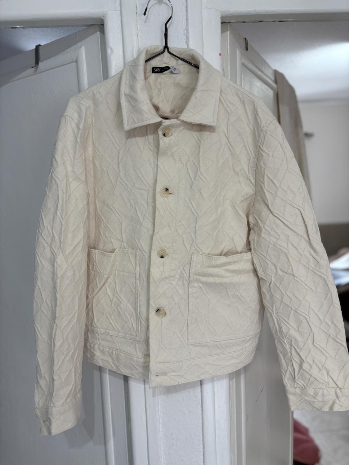 Beige padded jacket hanging on a coat hanger, with buttons down the front and large pockets, against a backdrop of white doors.