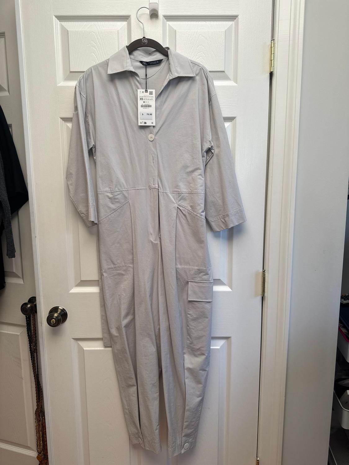 Light grey jumpsuit hanging on a white door, with visible price tag. Long-sleeved design with front pockets.