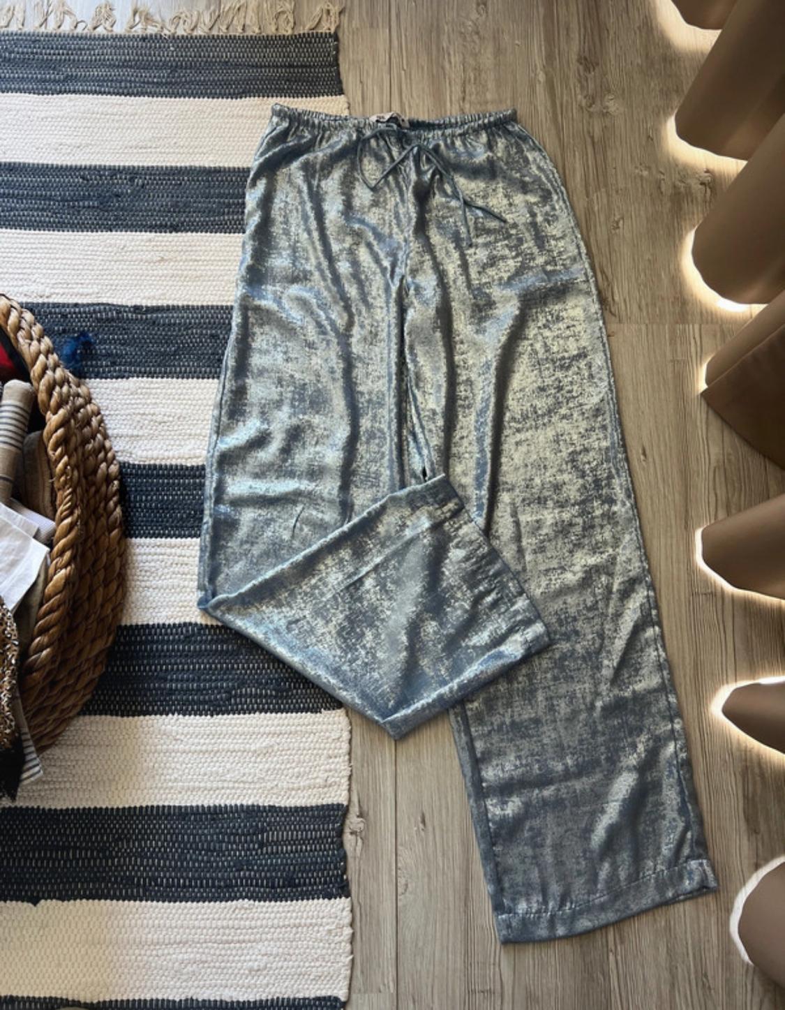 Metallic wide-leg trousers with drawstring, on a wooden floor and striped rug. Relaxed atmosphere with a wicker basket on the left.