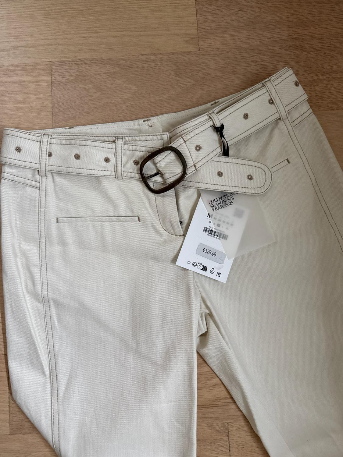 Beige trousers with a belt on a wooden floor. Text: COLLECTION SEASON: S/S YEAR: 2025 M XS $ 129.00.