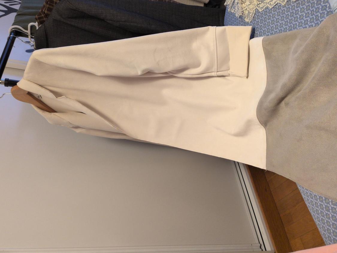 Beige coat hanging on a coat rack next to other dark garments. White wall background and wooden floor.