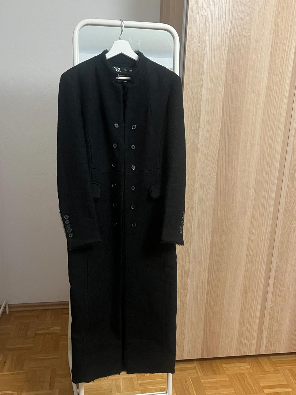 Long black coat hanging on a white coat rack in front of a mirror, with double-breasted buttons and a light wooden wall and door background.