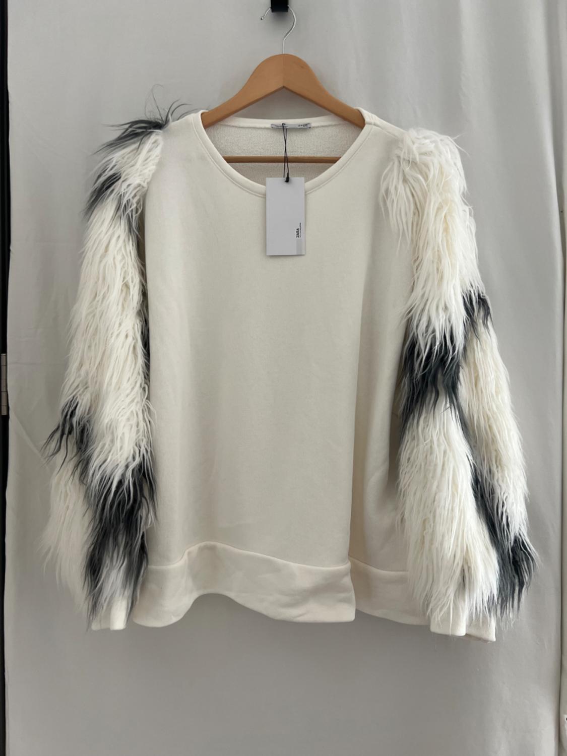 White sweatshirt hanging on a wooden hanger. Its long black and white faux fur sleeves stand out. Label with ZARA text. Light fabric background.