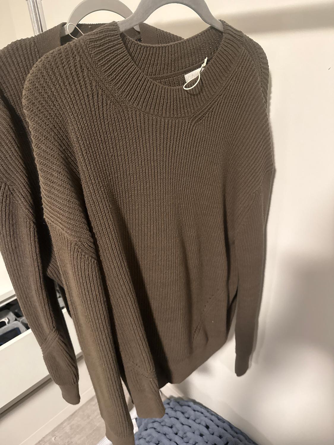 Chunky brown knitted jumper hanging on a coat hanger, reflected in a mirror. Background with drawers and a blue knitted blanket.