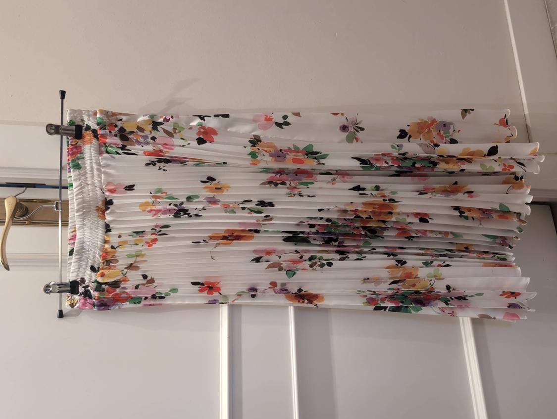 White pleated skirt with a multicoloured floral print and elasticated waist, hanging on a hanger over a white door.