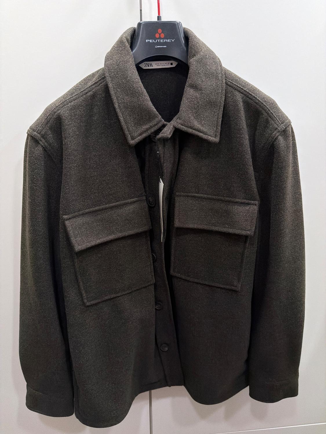 Dark grey jacket with large pockets and buttons, hanging on a black Peuterey branded hanger, against a white background.