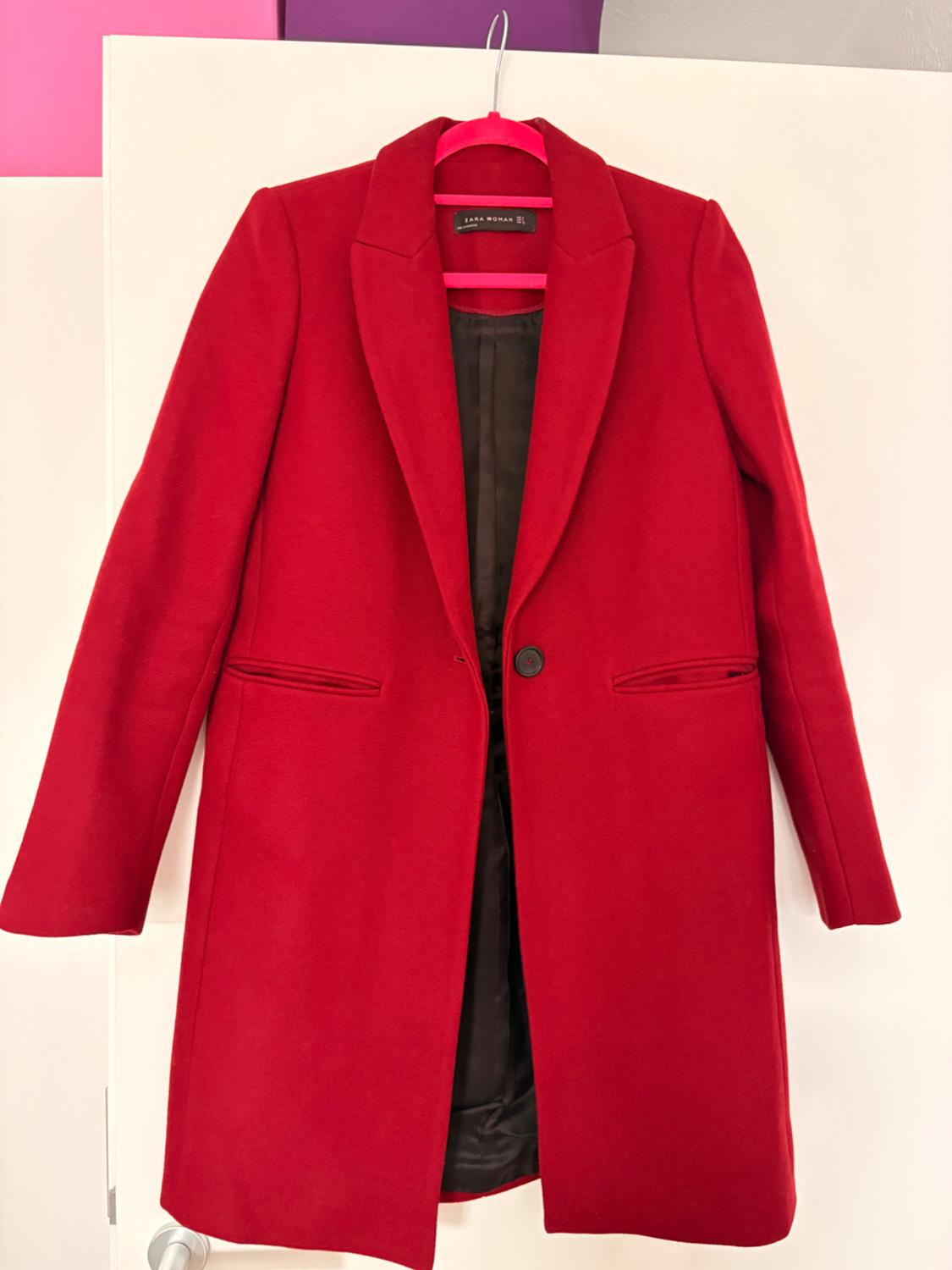 Red coat hanging on a pink hanger, with a background of pink and purple panels. Label visible on the inside.