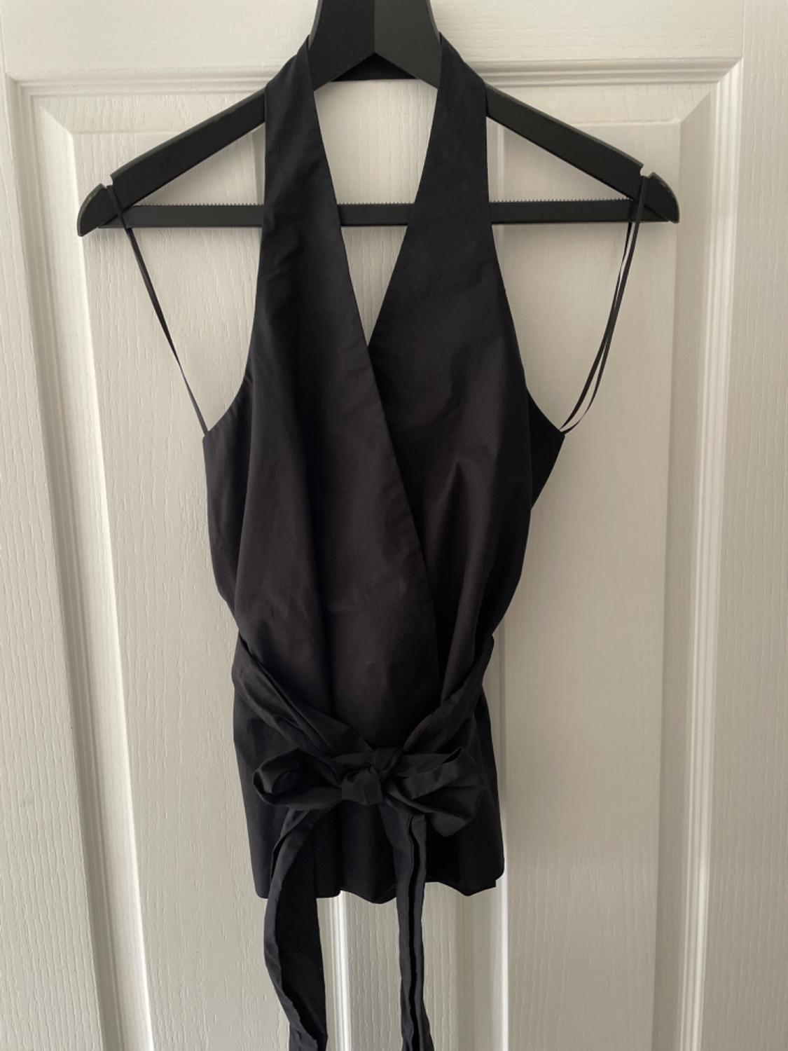 Black halter-neck crossover top with waist tie, hanging on a black hanger against a white door. Casual, relaxed style.