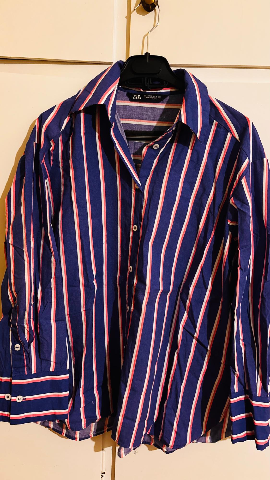 Vertical striped shirt in blue, red, and white, hanging on a black hanger against a light background.