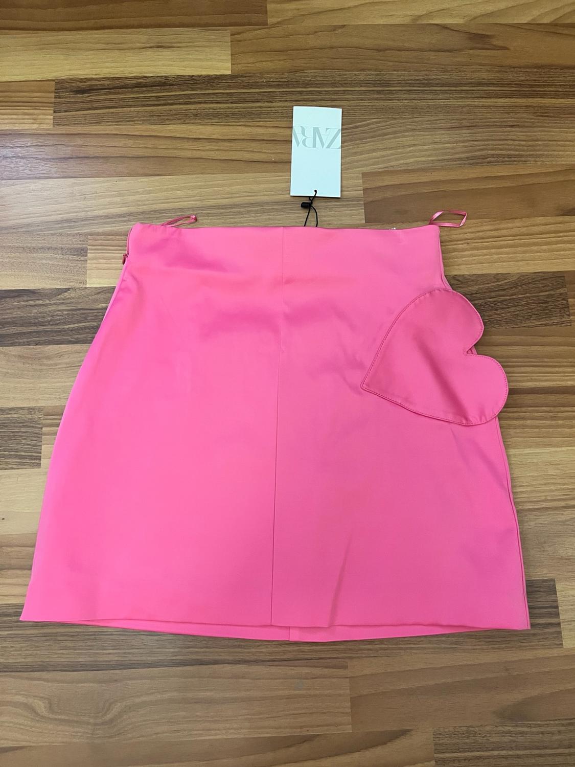 Pink miniskirt with a heart-shaped pocket, laid out on a wooden floor. Label visible with the text ZARA.