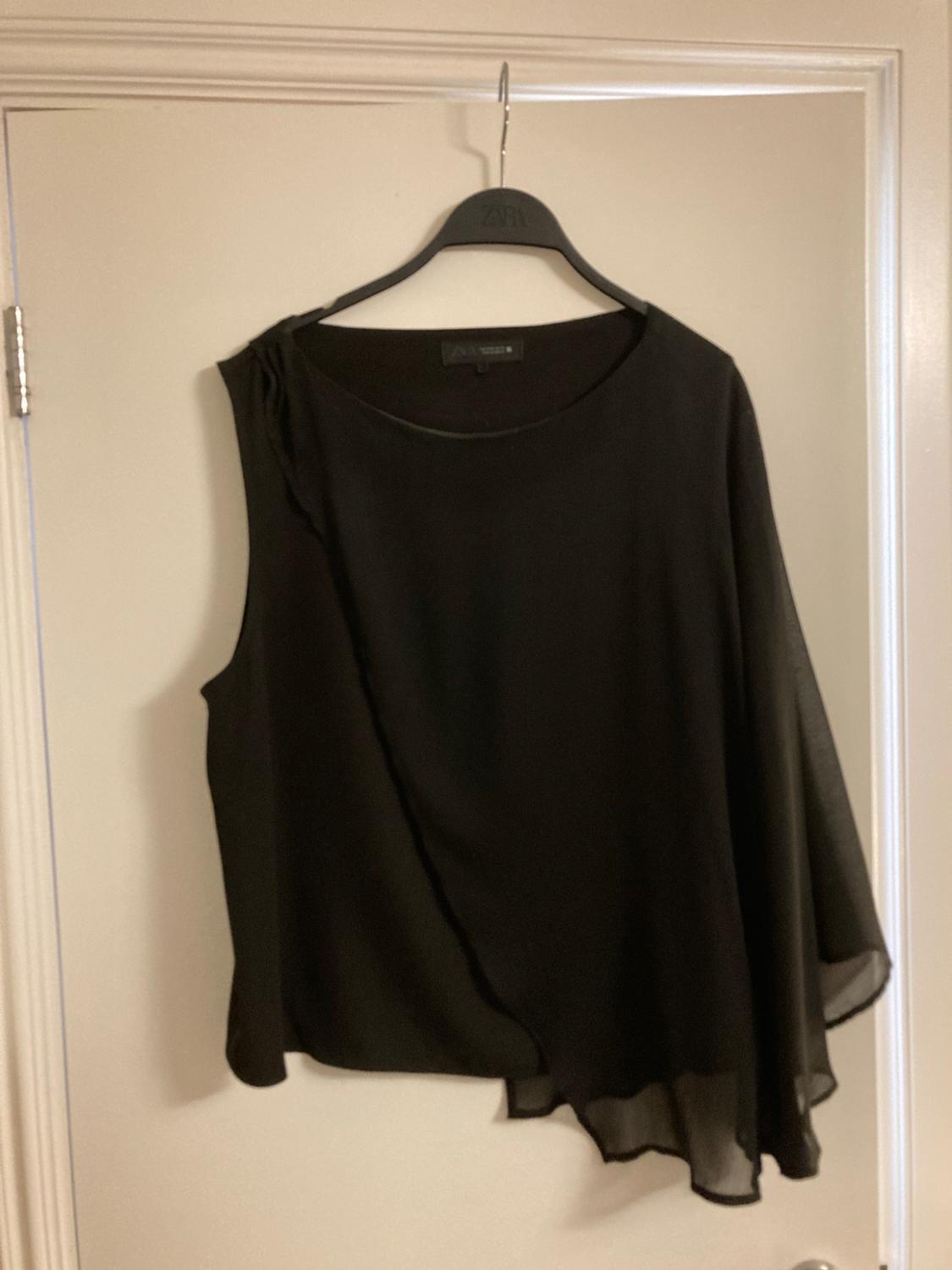 Black asymmetric flowing fabric top hanging on a hanger on a white door. You can read zara on the hanger and the label.