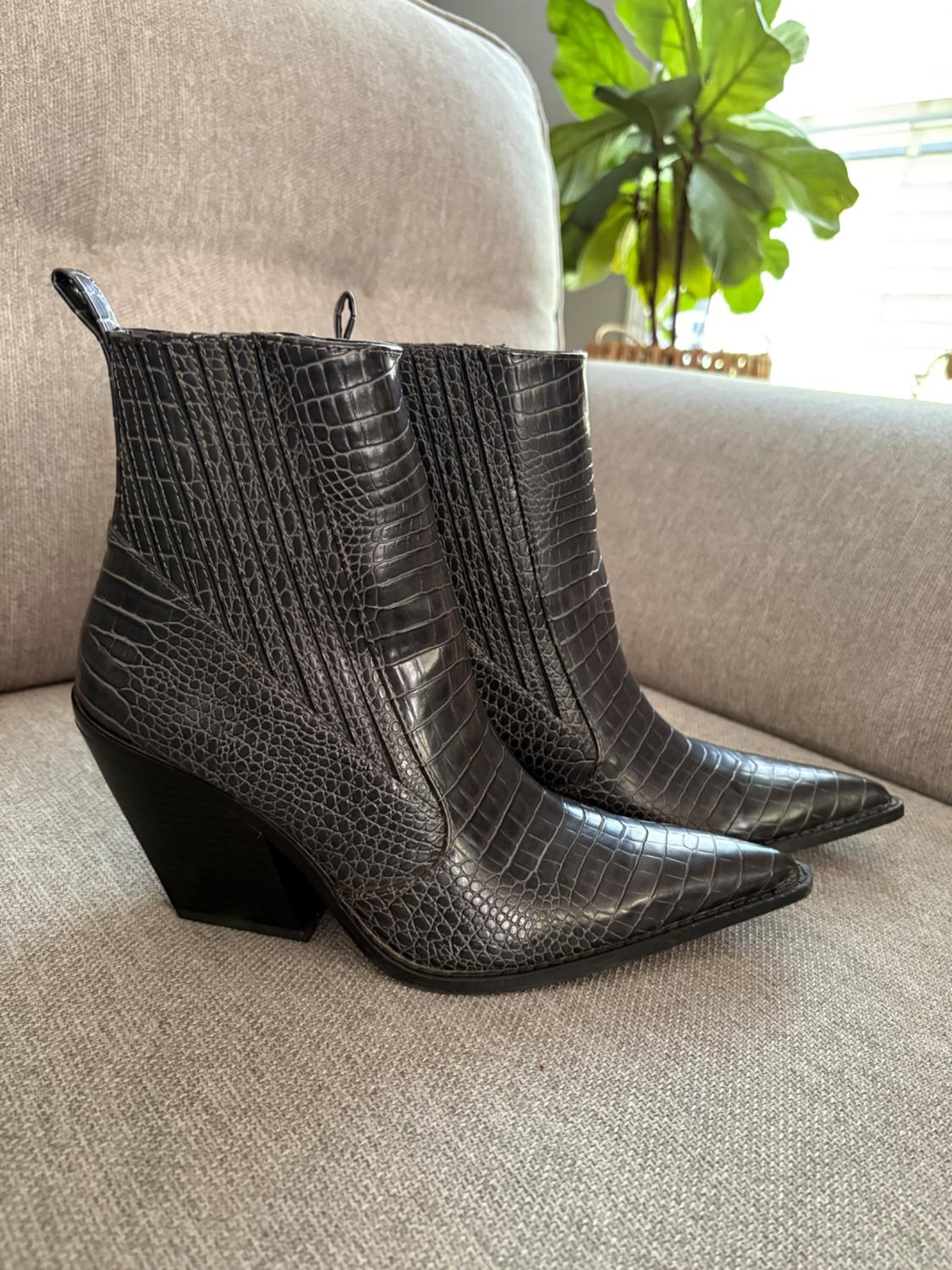 A pair of dark gray ankle boots with a crocodile-skin texture, a pointed toe, and a block heel, on a light gray fabric sofa.