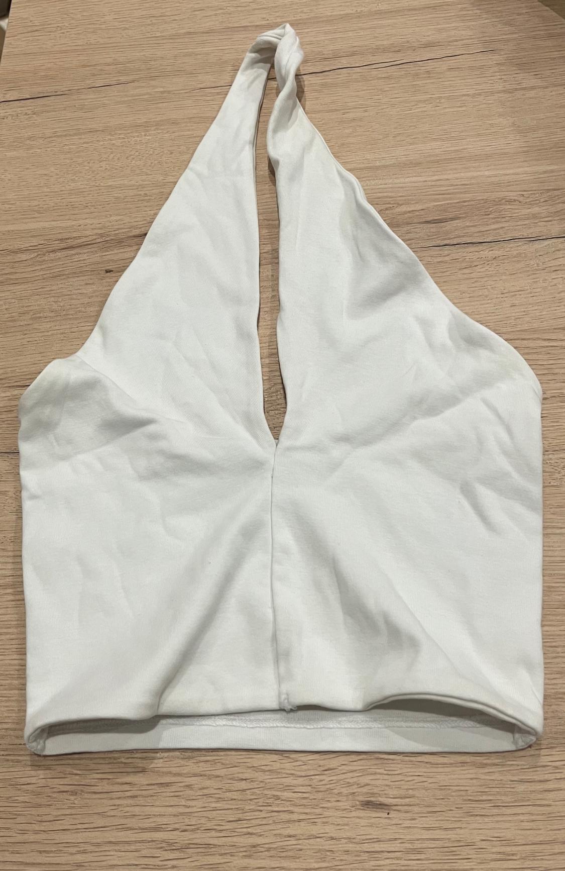 White halter neck crop top with a V-neckline and centre seam, laid out on a light wood surface. Casual style.