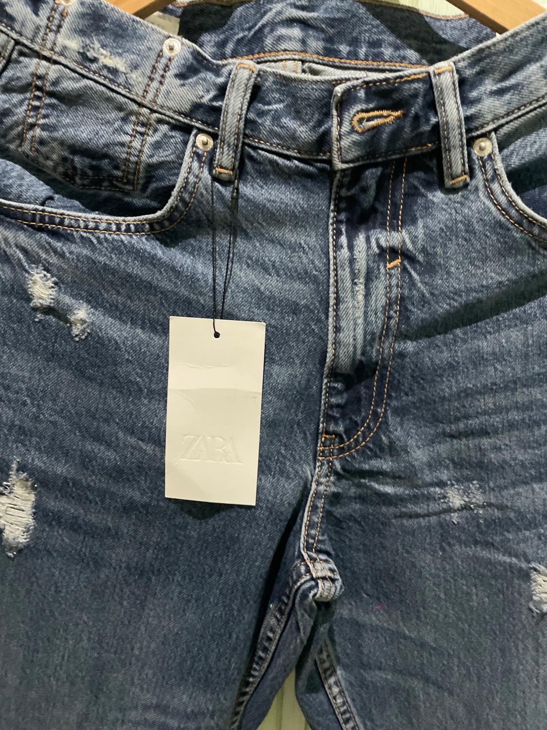 Front detail of distressed blue jeans on a wooden hanger. White hanging tag with ZARA text.