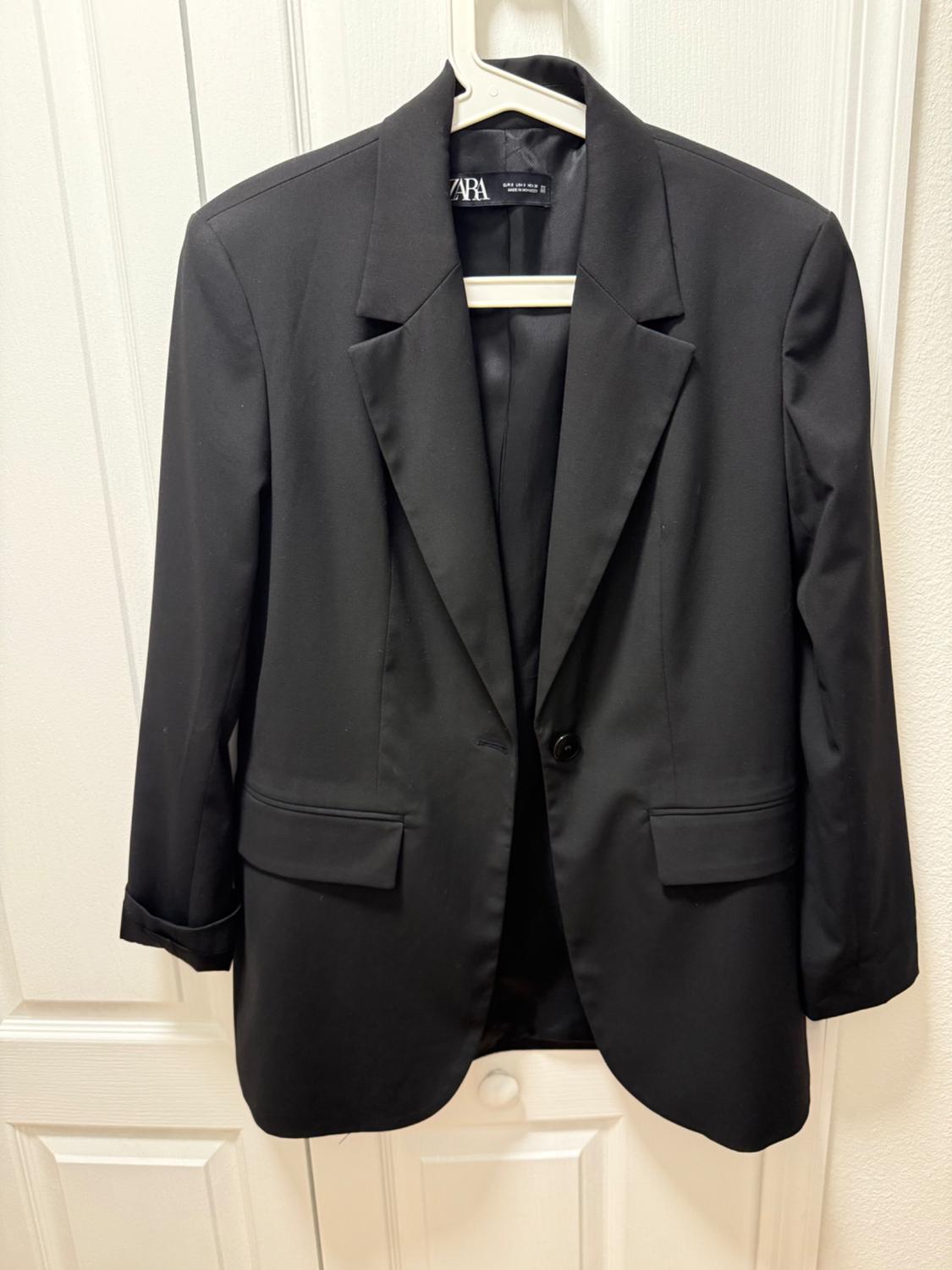 Black jacket hanging on a white hanger in front of a white door. Visible tag with the brand "ZARA".