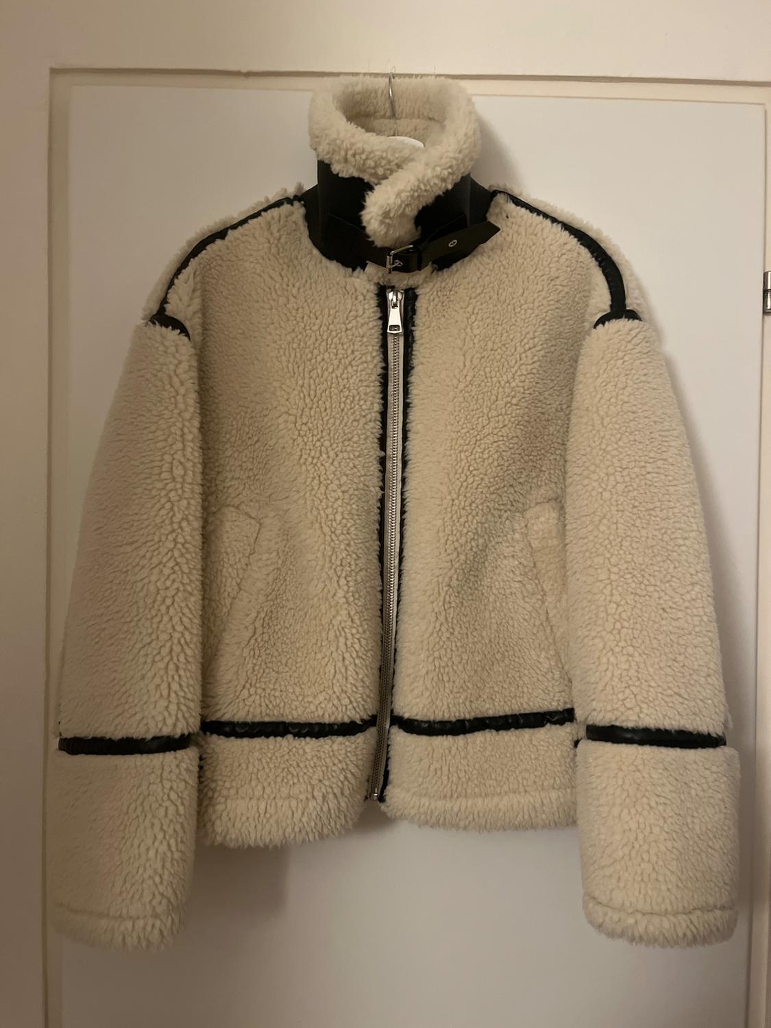 Beige fleece jacket with black details and a front zip, hanging on a white door.