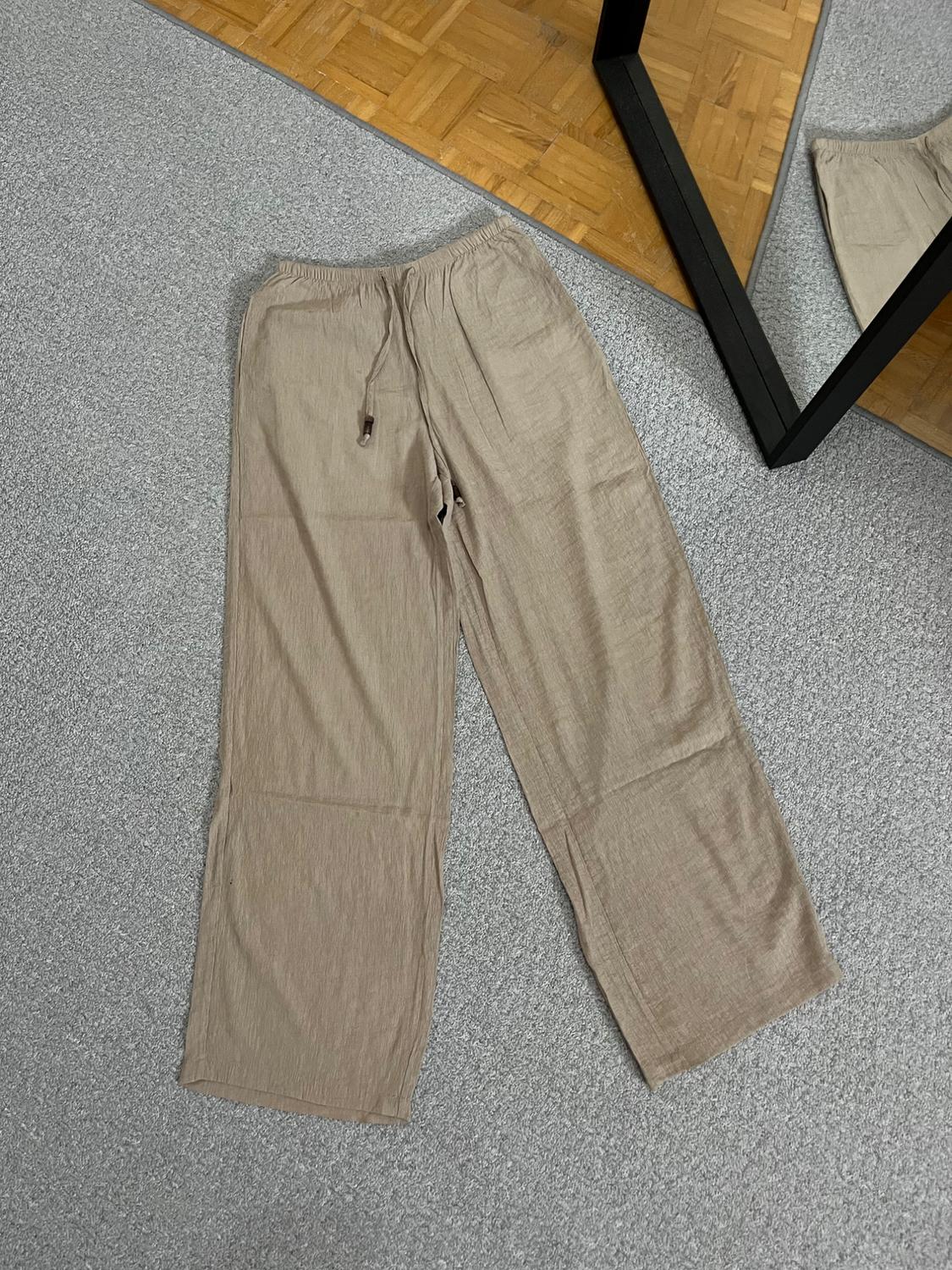 Beige wide-leg trousers with an elasticated waistband and drawstring, on a grey rug next to a wooden floor and a mirror.