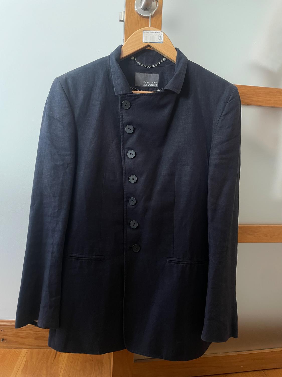Dark blue jacket with an asymmetrical button fastening hanging on a wooden hanger. ZARA MAN text on the label. White background.