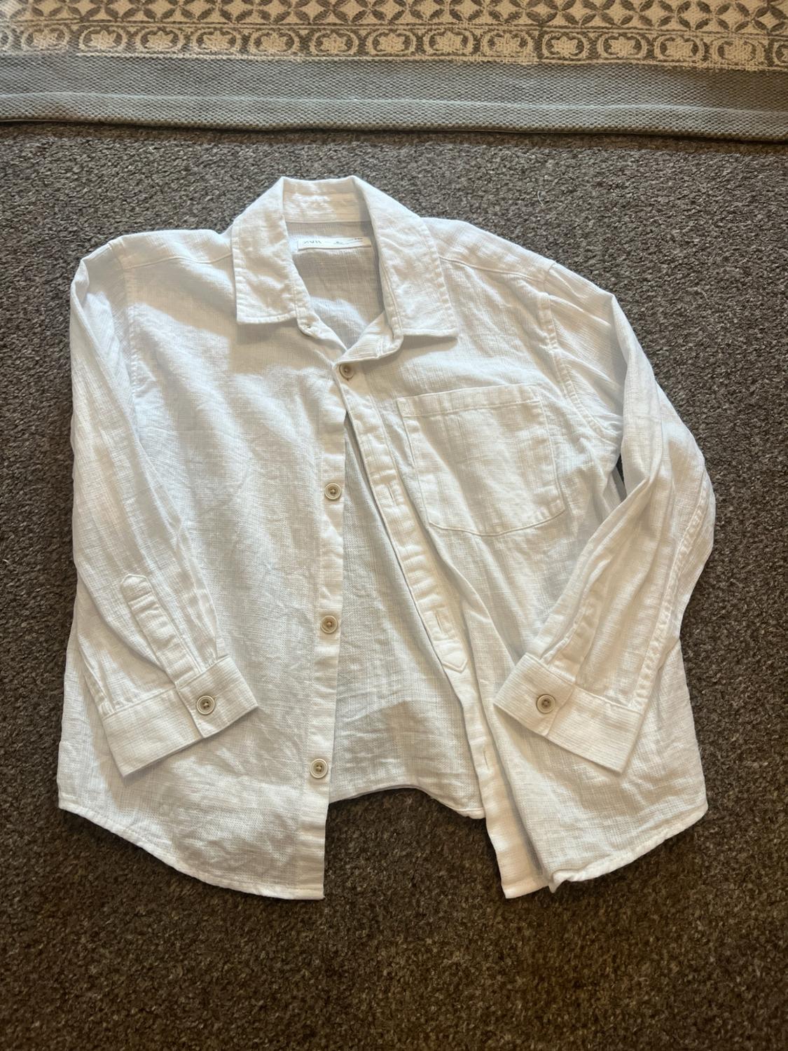 Textured long-sleeved white shirt with buttons and a pocket, laid out on a brown carpet. ZARA label on the collar.