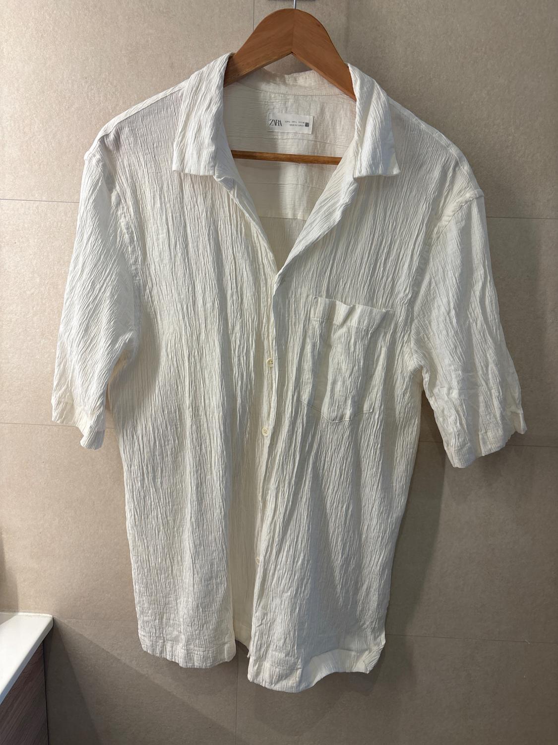 Short-sleeved white shirt, crinkled texture and pocket, on a wooden hanger against a beige tiled wall. Label with text: ZARA.