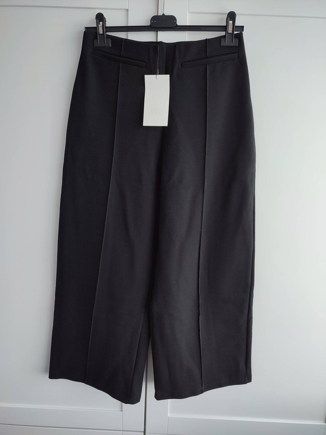 Black wide-leg trousers with front seam, hanging on a hanger against a white background. It has a white label.