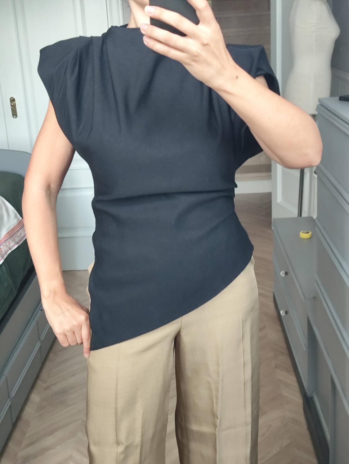 Mirror selfie of a woman in a room. She is wearing a black top with shoulder pads and an asymmetric hem, and wide-leg beige trousers.