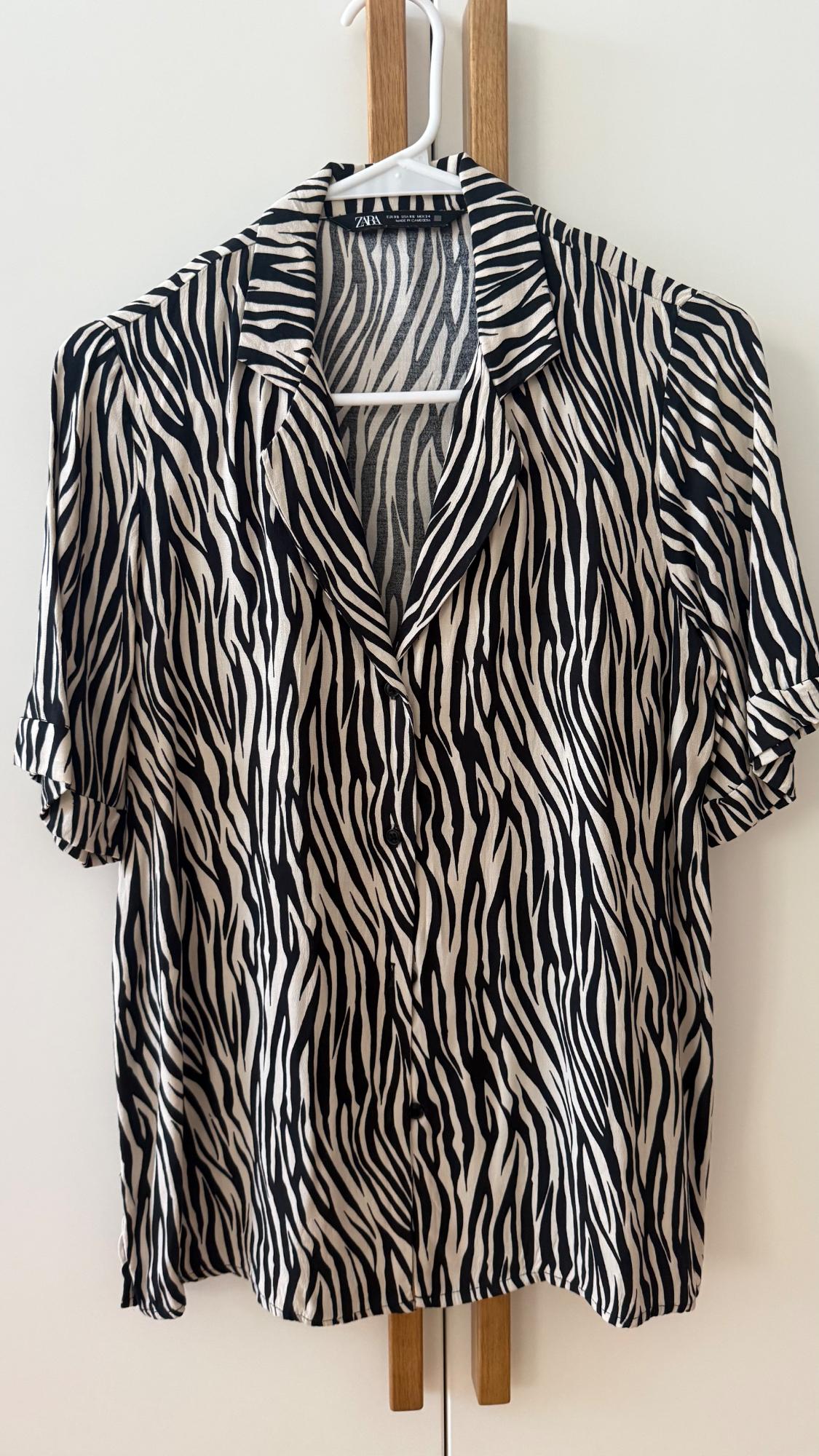 Short-sleeved zebra print shirt hanging on a white hanger over doors with wooden handles. Text: ZARA.