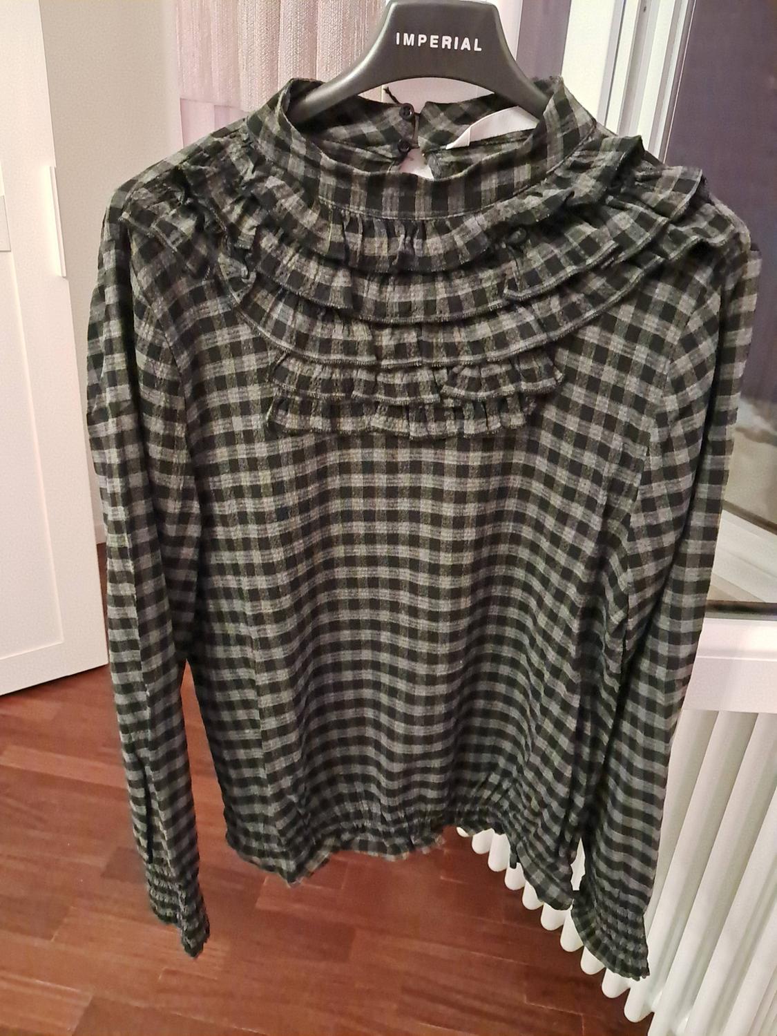 Checked blouse with front ruffles, hanging on a hanger bearing the text IMPERIAL. Interior background with wooden flooring.