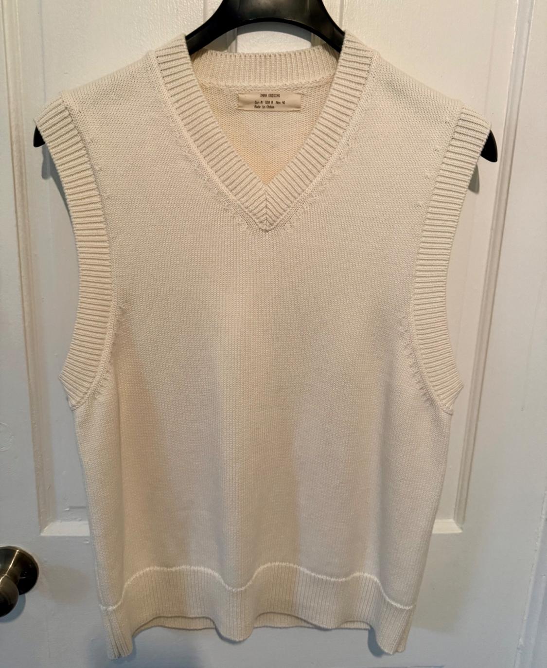Cream V-neck sleeveless vest, hanging on a black hanger in front of a white door. Label visible on the collar.