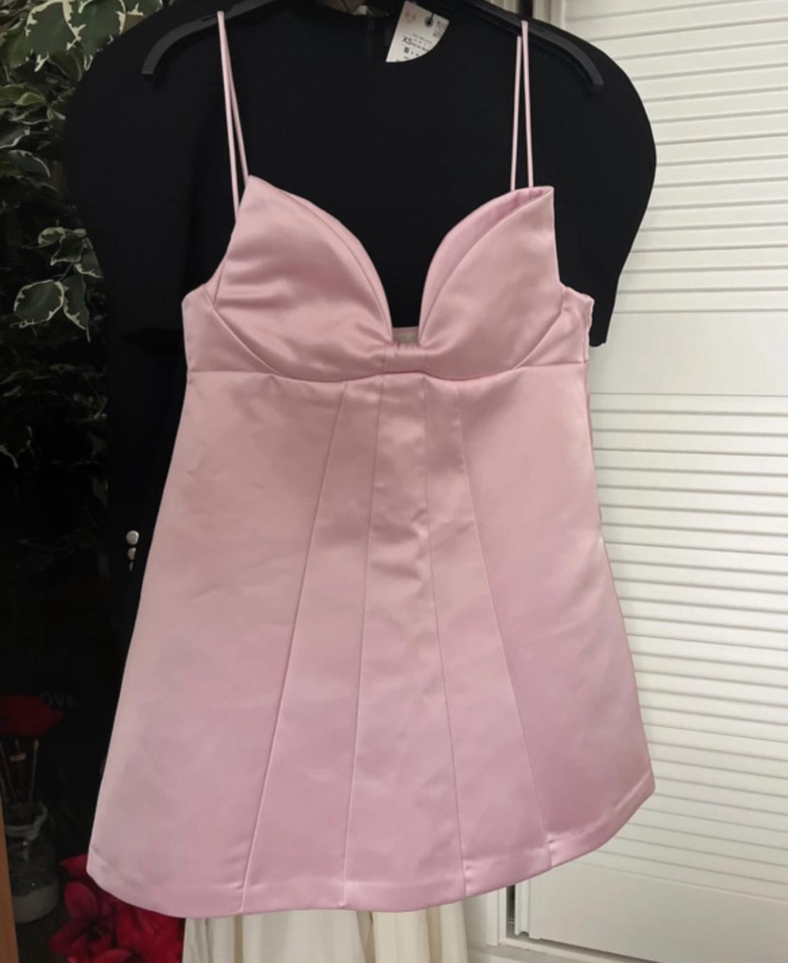 Short satin pink V-neck cami dress hung over a black garment. Background with white door and LOVE text. XS label.