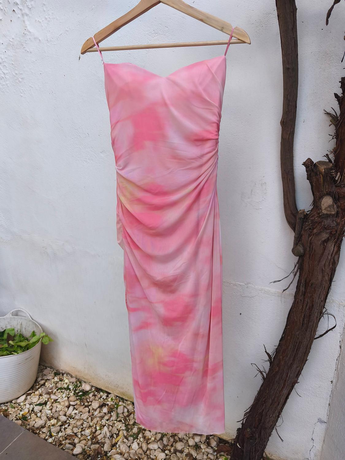 Long, thin-strapped dress in pink and yellow tones, hanging on a wooden hanger against a white wall.