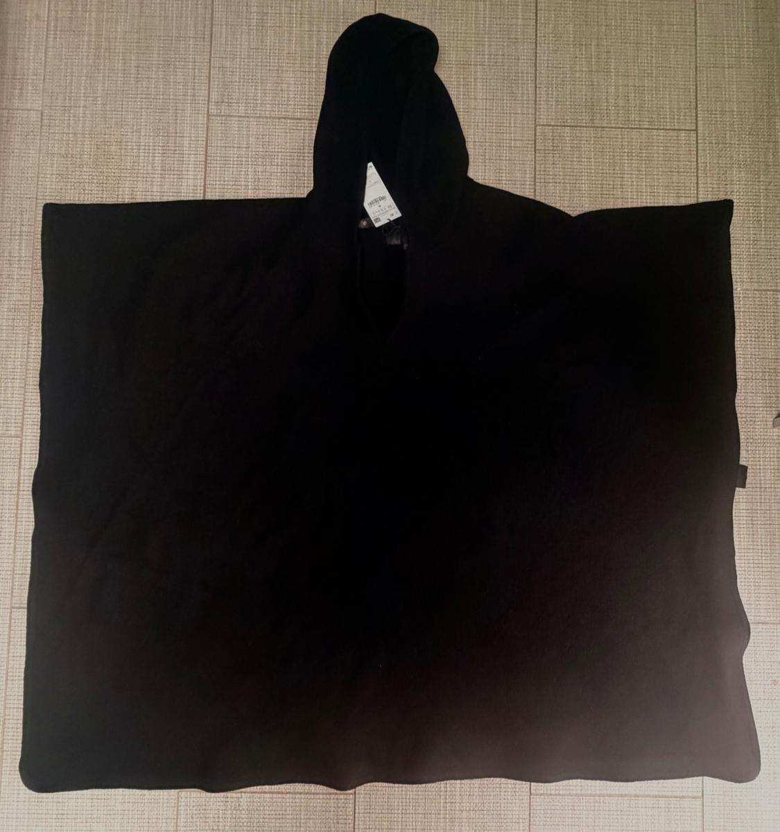 Black cape with an extended hood laid out on a light wooden floor. Label visible on the inside of the hood.