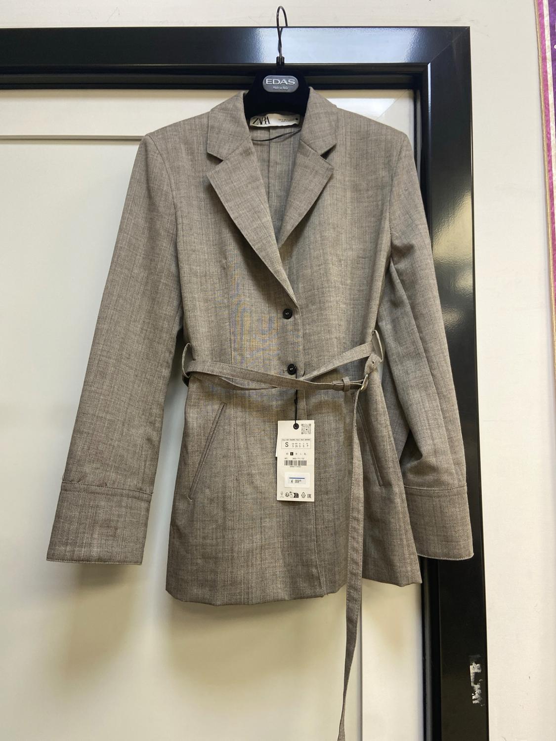 Grey blazer with a belt and black buttons hanging on a hanger. Text: EDAS, ZARA, S.
