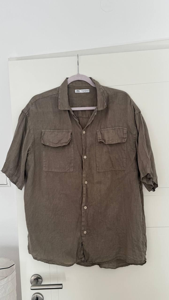 Olive green short-sleeved shirt with buttons and two pockets, hanging on a hanger on a white door. ZARA text on the label.