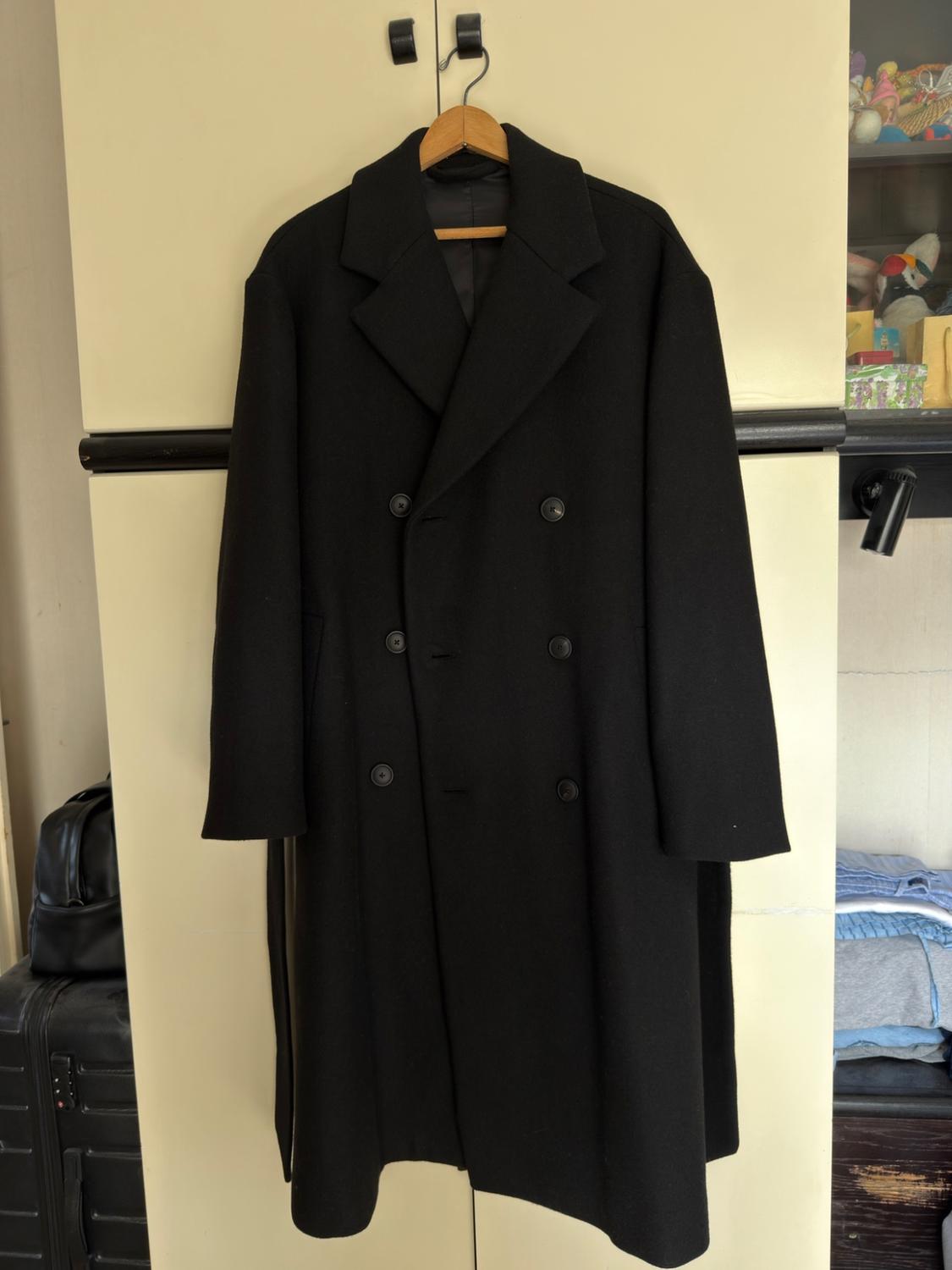 Long black double-breasted coat hanging on a wooden hanger on a light wardrobe in a domestic setting.