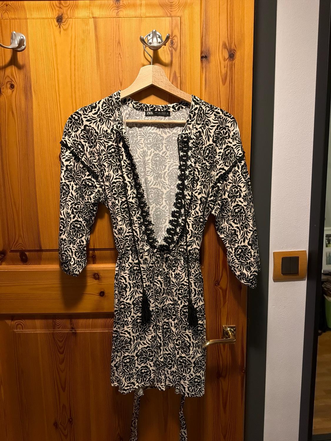 Short dress with a black and white floral print, V-neckline with lace and tassels. Hanging on a wooden door. ZARA label.