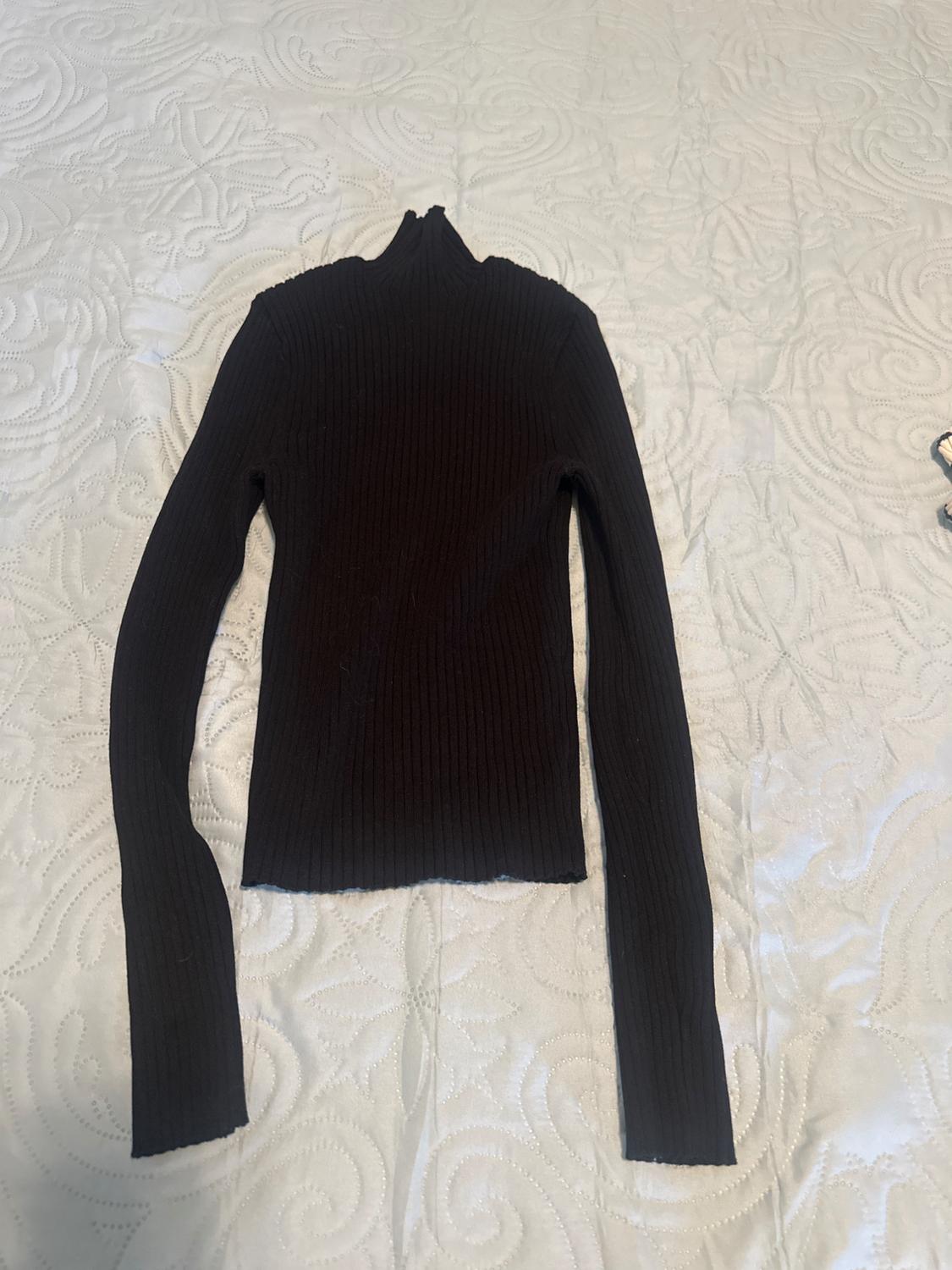 Black ribbed knit jumper, with a high neck and long sleeves, laid out on a white bedspread with a curved pattern texture.