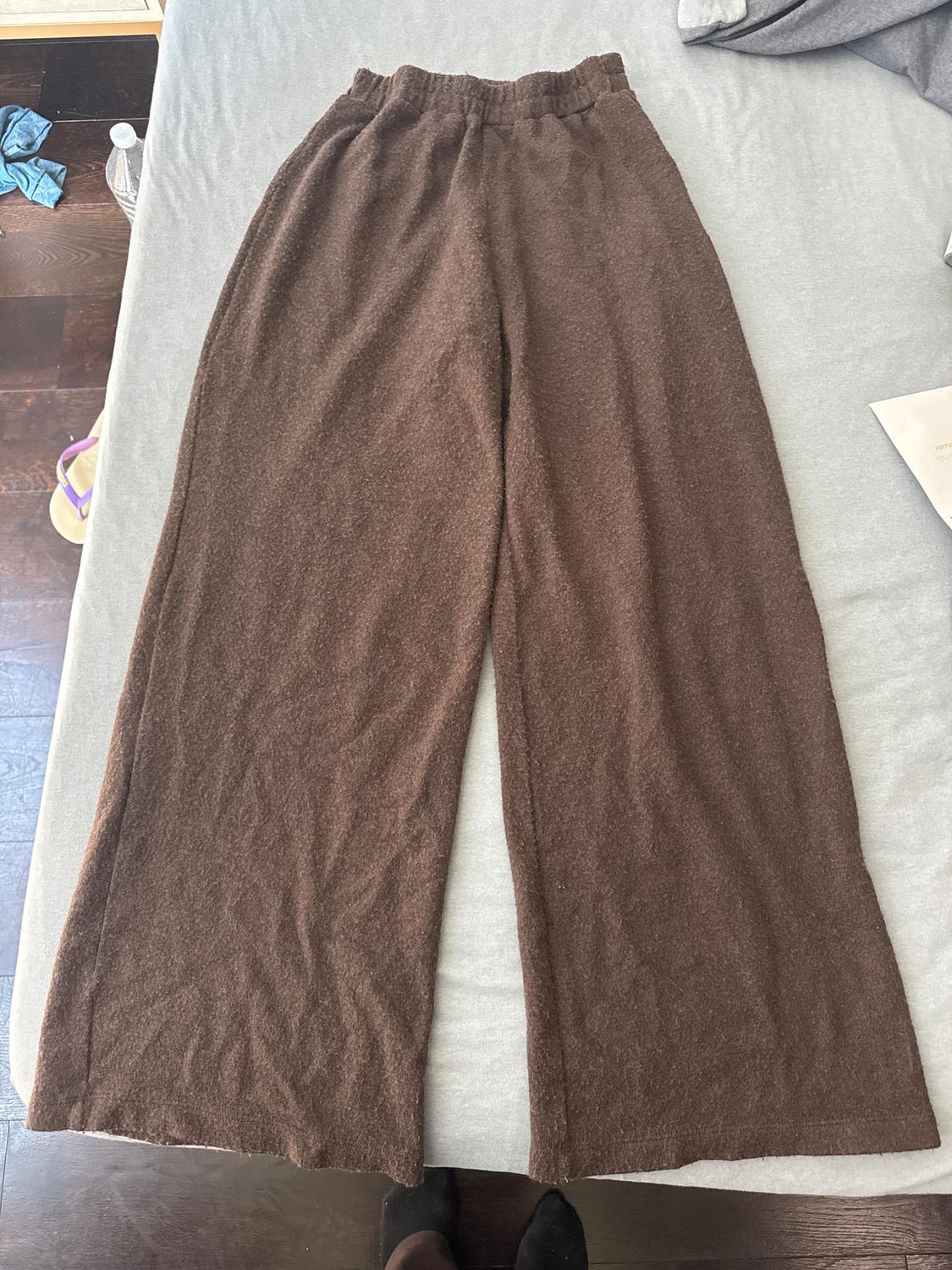 Brown wide-leg trousers on a grey bed. In the background, a water bottle and sandals on the wooden floor.