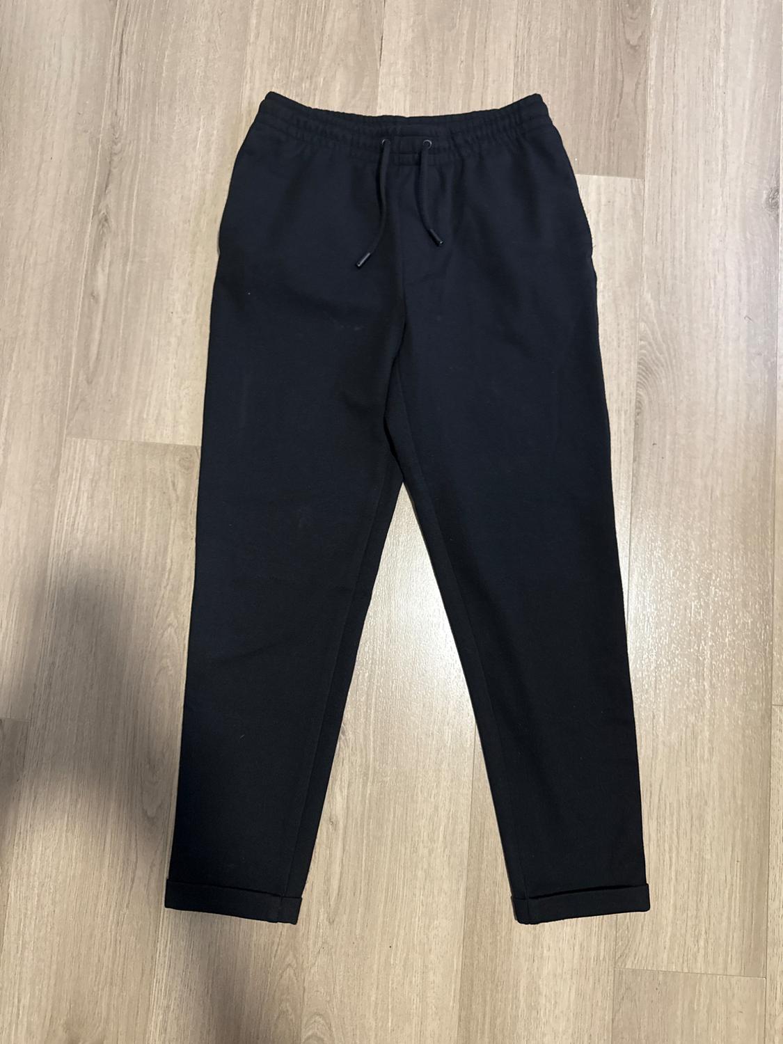 Casual black trousers with an elasticated waist, drawstring and turn-up hems, laid out on a light wood floor in a relaxed composition.