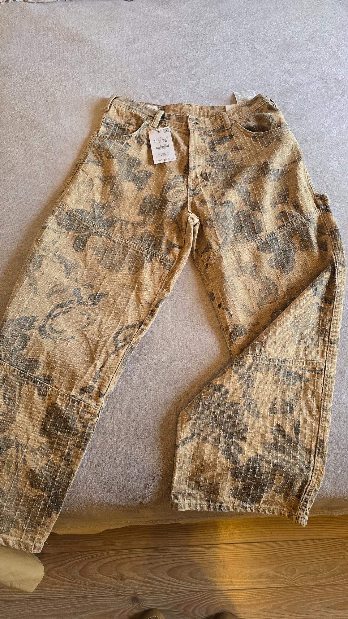 Denim trousers with a camouflage print in brown and grey tones, placed on a bed. Visible label on the waistband.