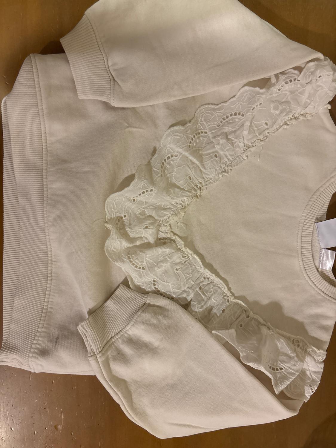 White sweatshirt with lace ruffles on the chest, long sleeves and ribbed cuffs, on a wooden surface.