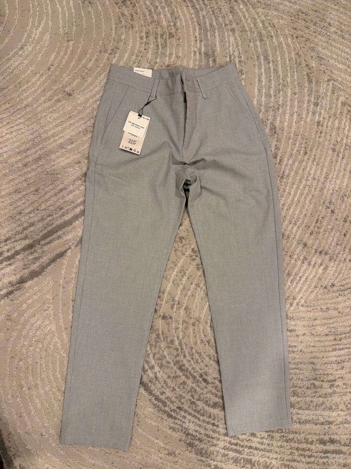 Light grey trousers laid out on a textured rug. A label bearing the text THE MELANGE PANT stands out.