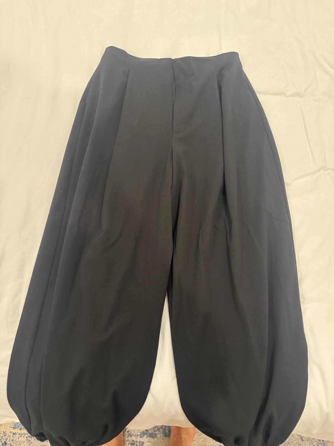 Wide black trousers with a gathered hem on a white sheet. A pair of bare feet protrudes from the lower edge, resting on a rug.