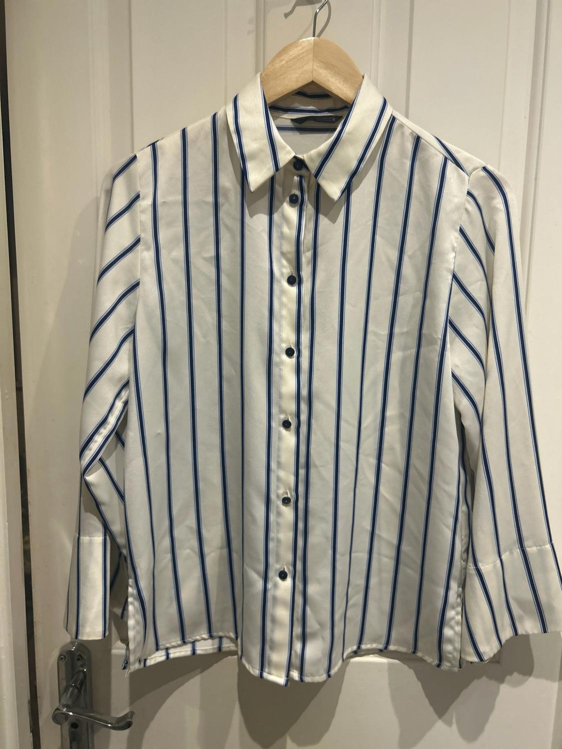 White long-sleeved shirt with blue vertical stripes and buttons, hanging on a wooden hanger in front of a white door.