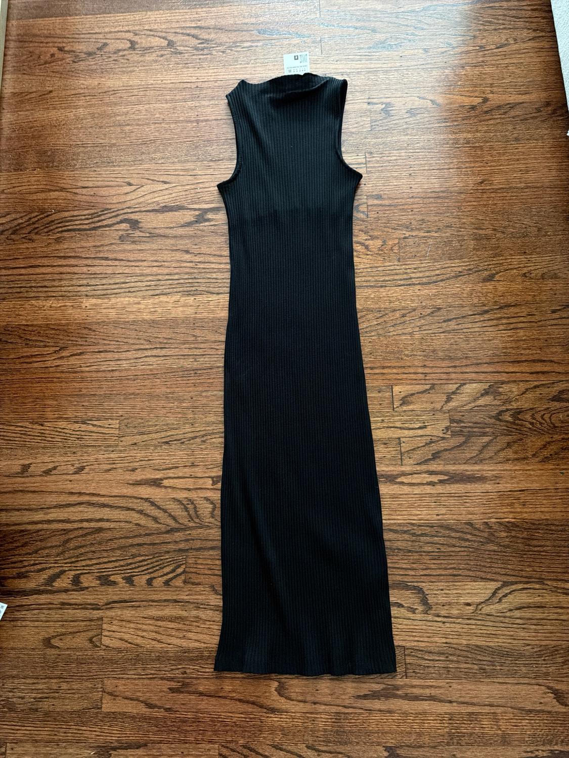 Black long sleeveless dress in ribbed knit with a high neck, on a wooden floor, with a price tag on the top.
