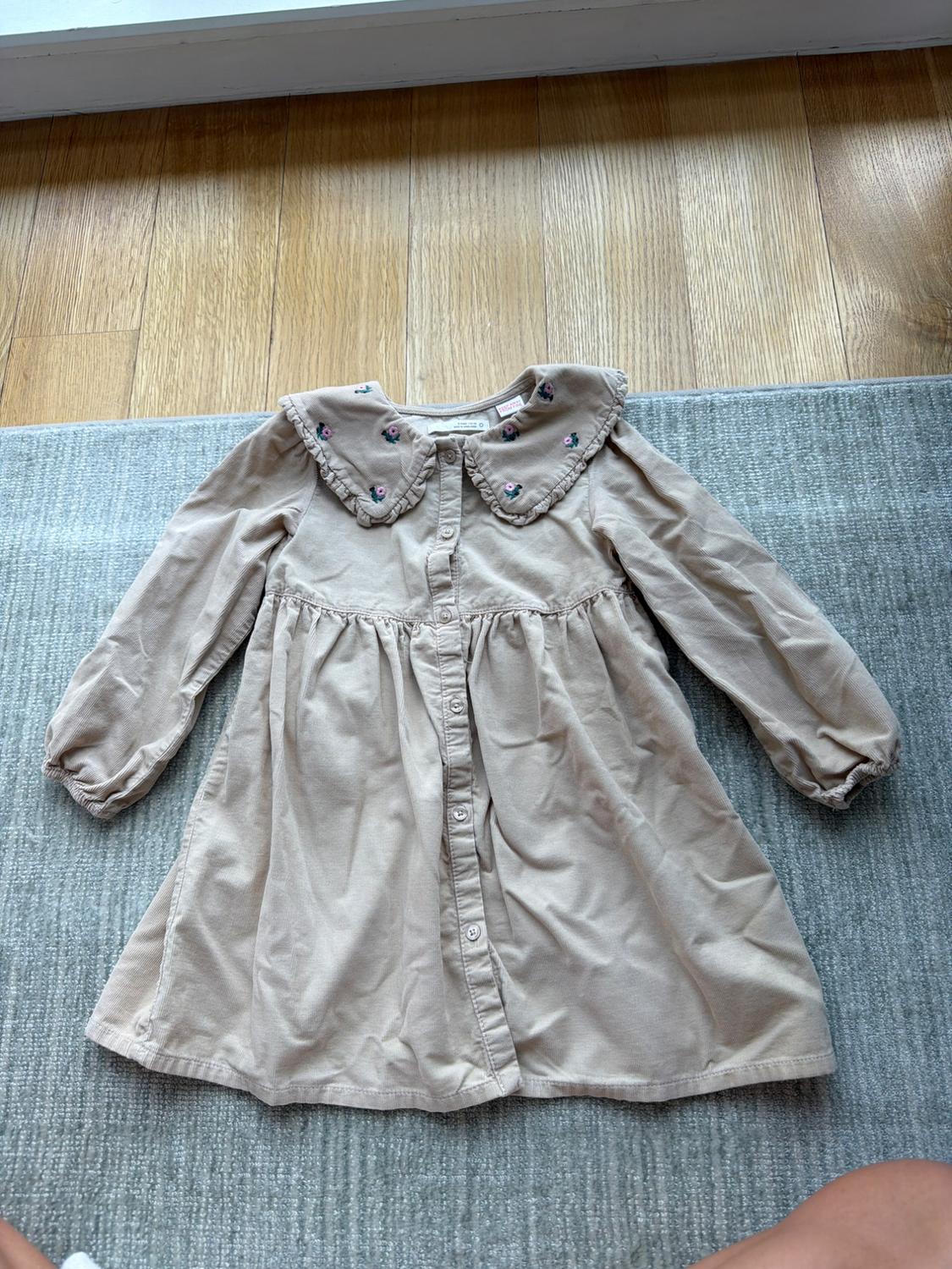 Long-sleeved beige dress with a collar embroidered with small flowers, on a grey rug and wooden floor.