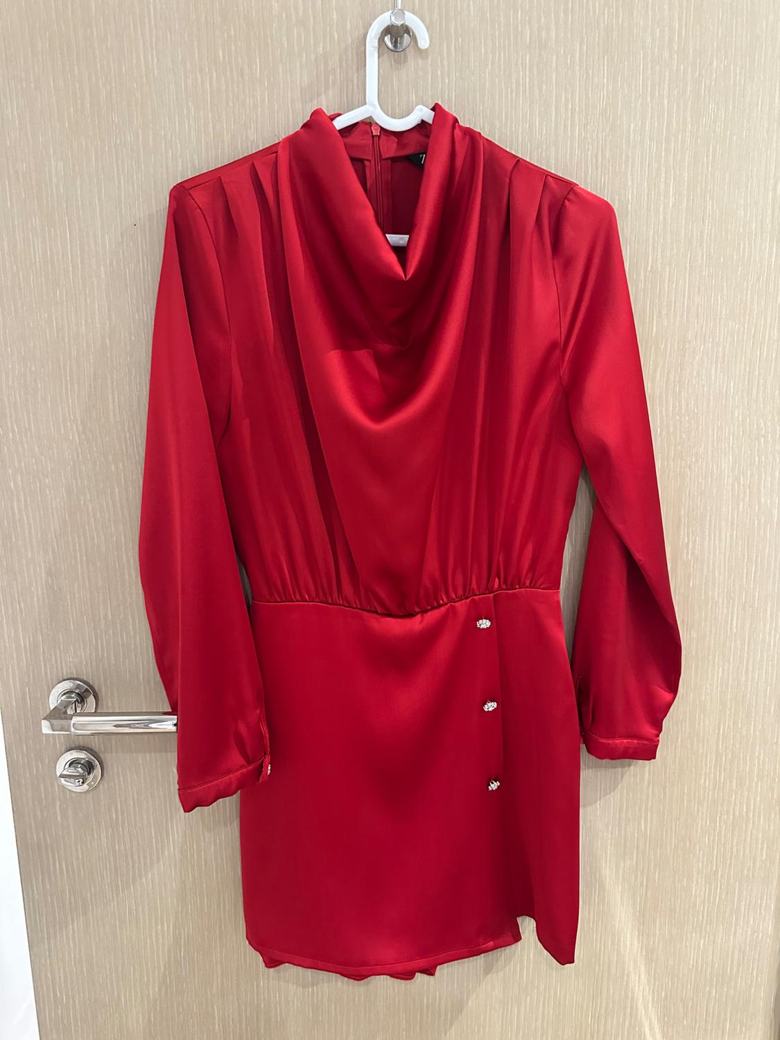 Long-sleeved red satin dress with a cowl neck and buttons on the skirt, hanging from a white hanger on a light wood door.
