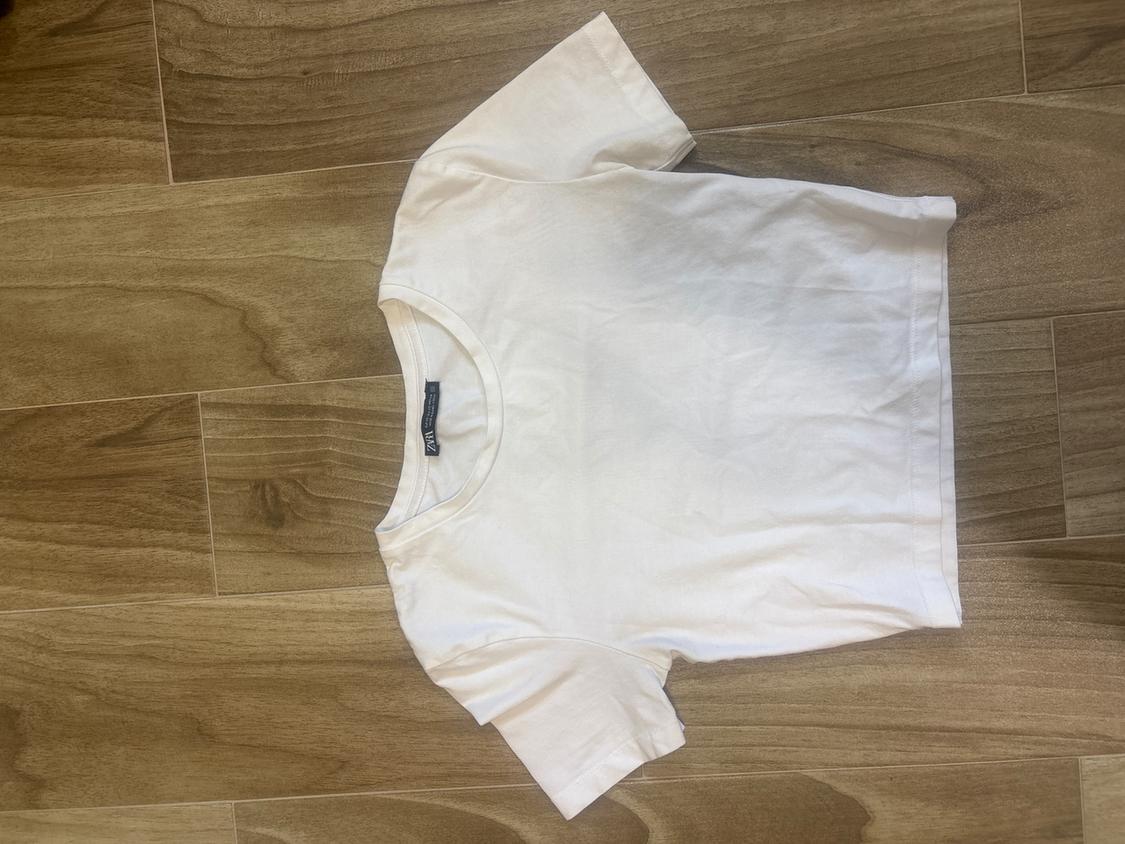 White short-sleeved t-shirt on a light wooden floor. Visible tag on the collar.