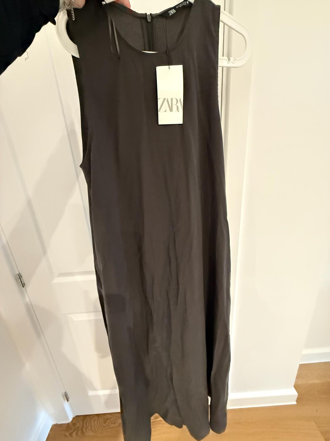 Person holding a dark grey sleeveless maxi dress on a white hanger in front of a door. Label with the text ZARA.
