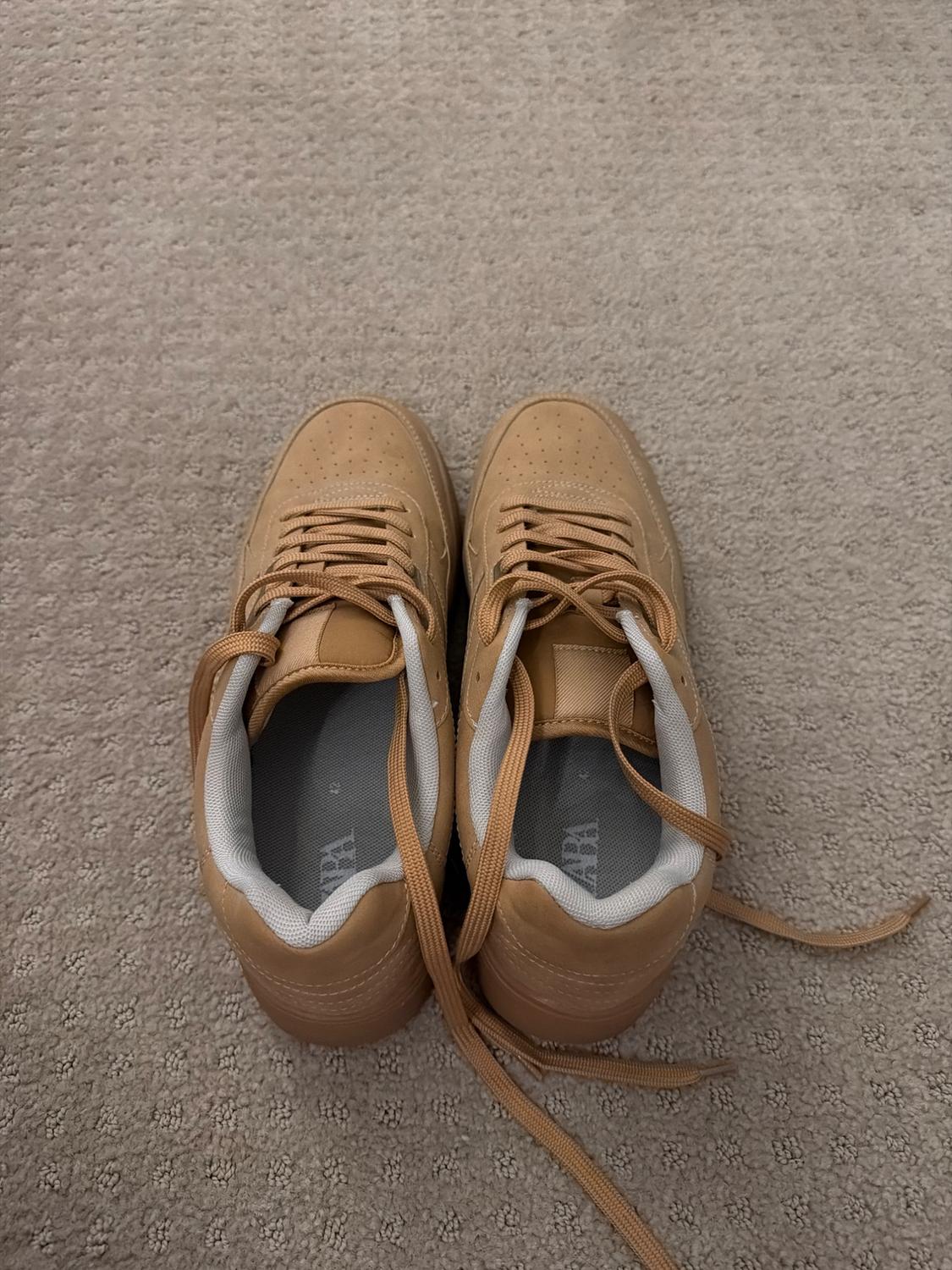 A pair of camel-colored sneakers on a beige rug. They have laces in the same tone and the ZARA logo visible on the inner insole.